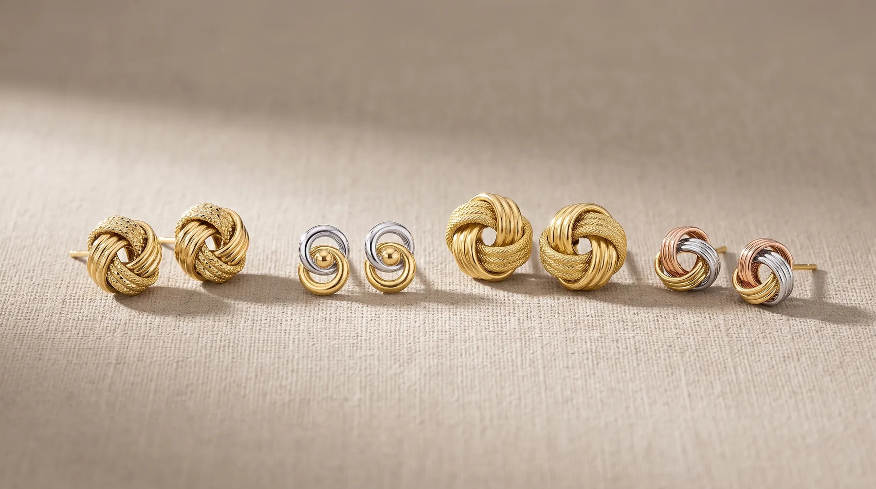 Eight pairs of knot-shaped stud earrings in gold and two-tone metal finishes arranged in a row on a neutral fabric surface.