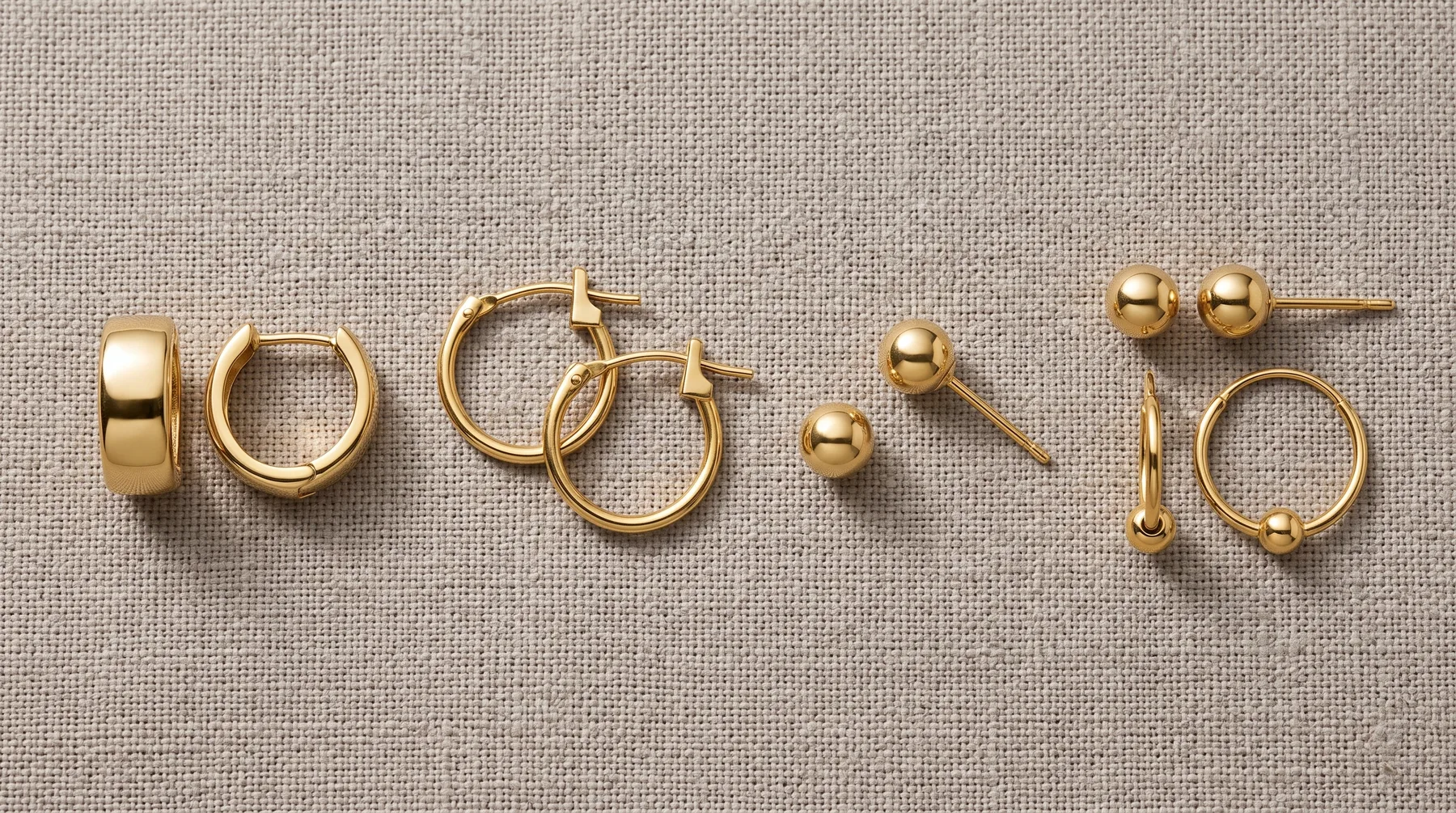 Six gold-toned jewelry pieces arranged on a beige linen background: a polished band ring, hoop earrings, chunky hoop earrings, a ball stud earring, a bar stud earring, ball stud earrings, and a textured ring with ball accents.