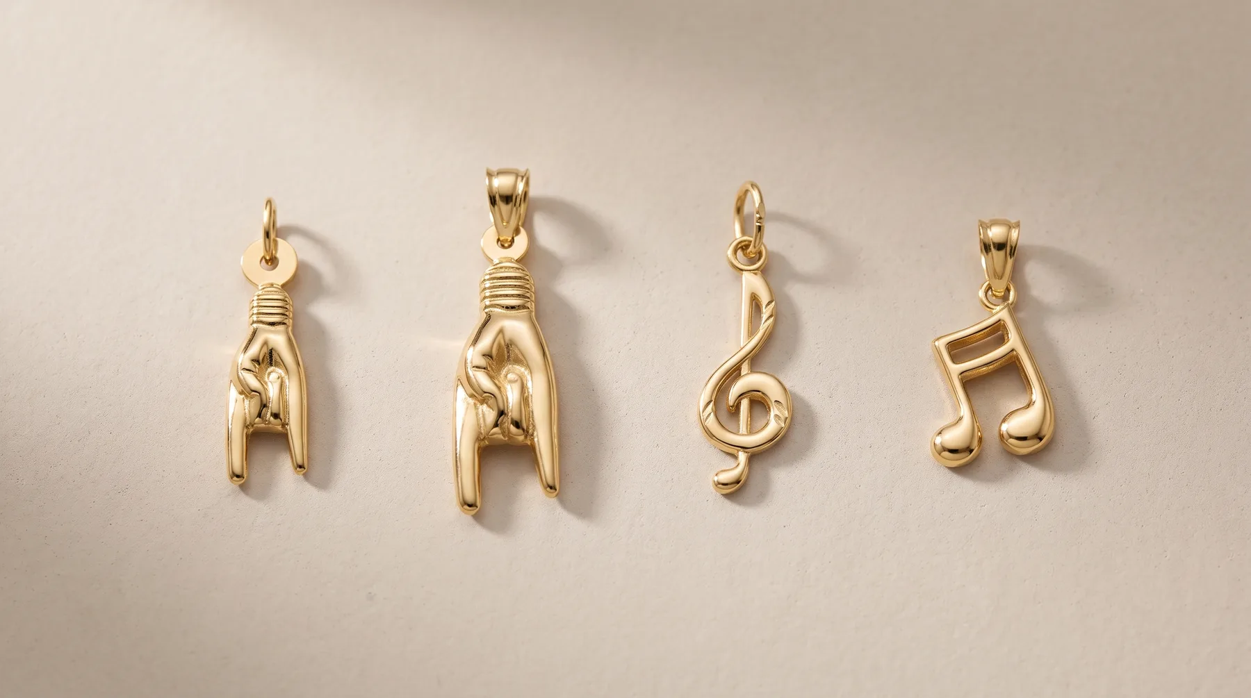 Four gold pendant charms displayed on a neutral background: a wishbone, a safety pin, a musical note, and a pair of eighth notes.
