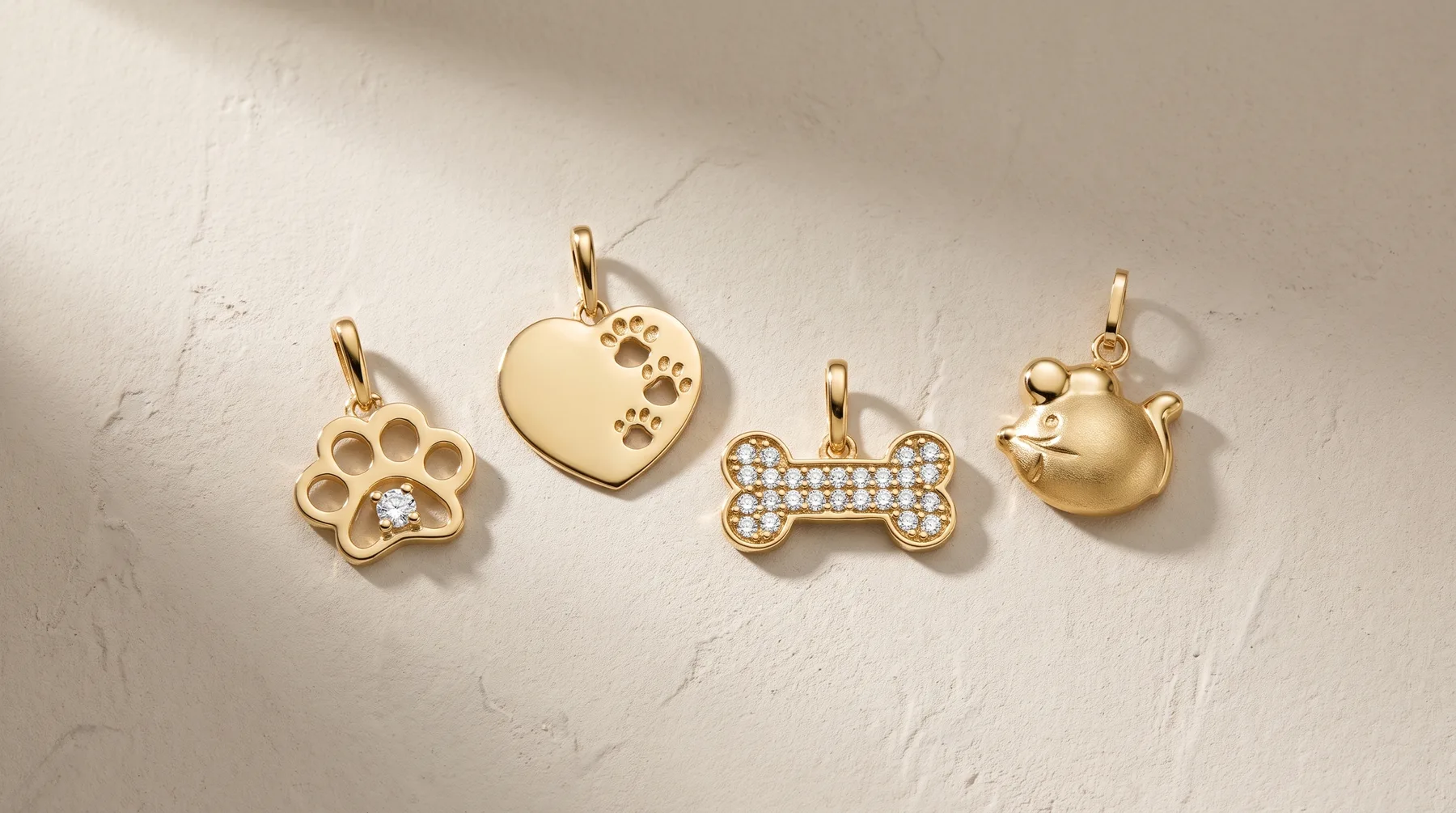 Four gold pet charms hang from a textured white surface: a paw print with a crystal accent, a heart, a bone studded with crystals, and a bell.