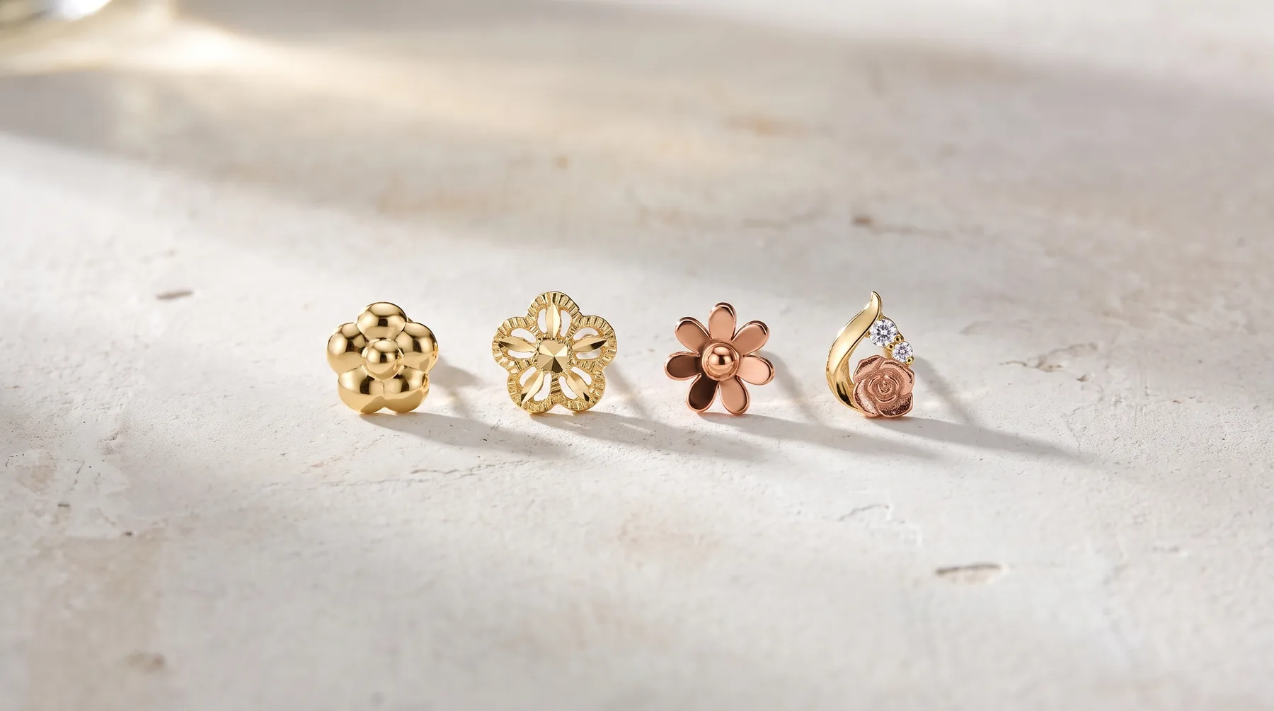 Four gold earring studs with different flower designs arranged in a row on a white textured surface.
