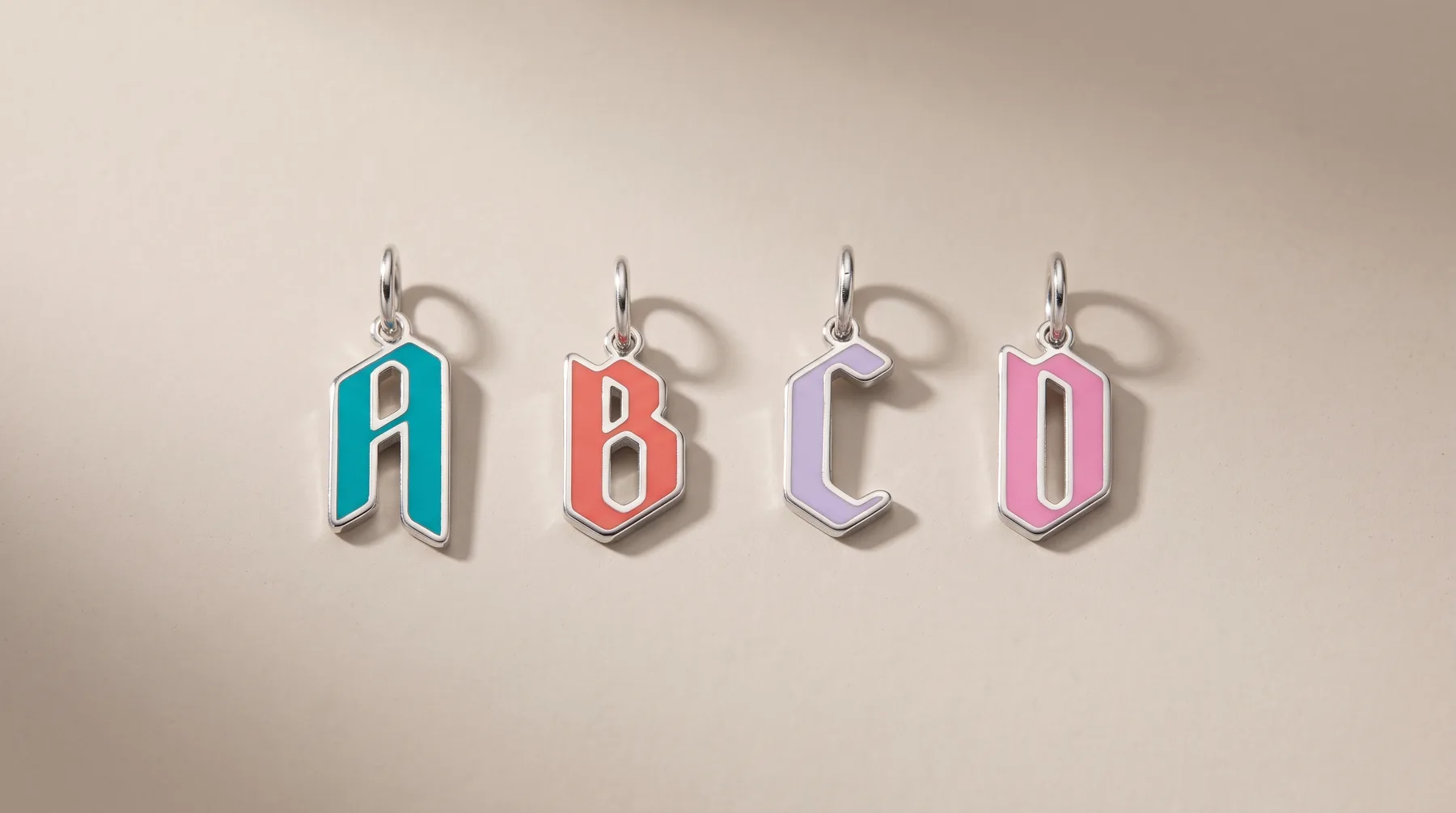Four colorful enamel letter charms spelling "ABCD" in teal, coral, purple, and pink, each with a silver loop attached, displayed against a neutral beige background.