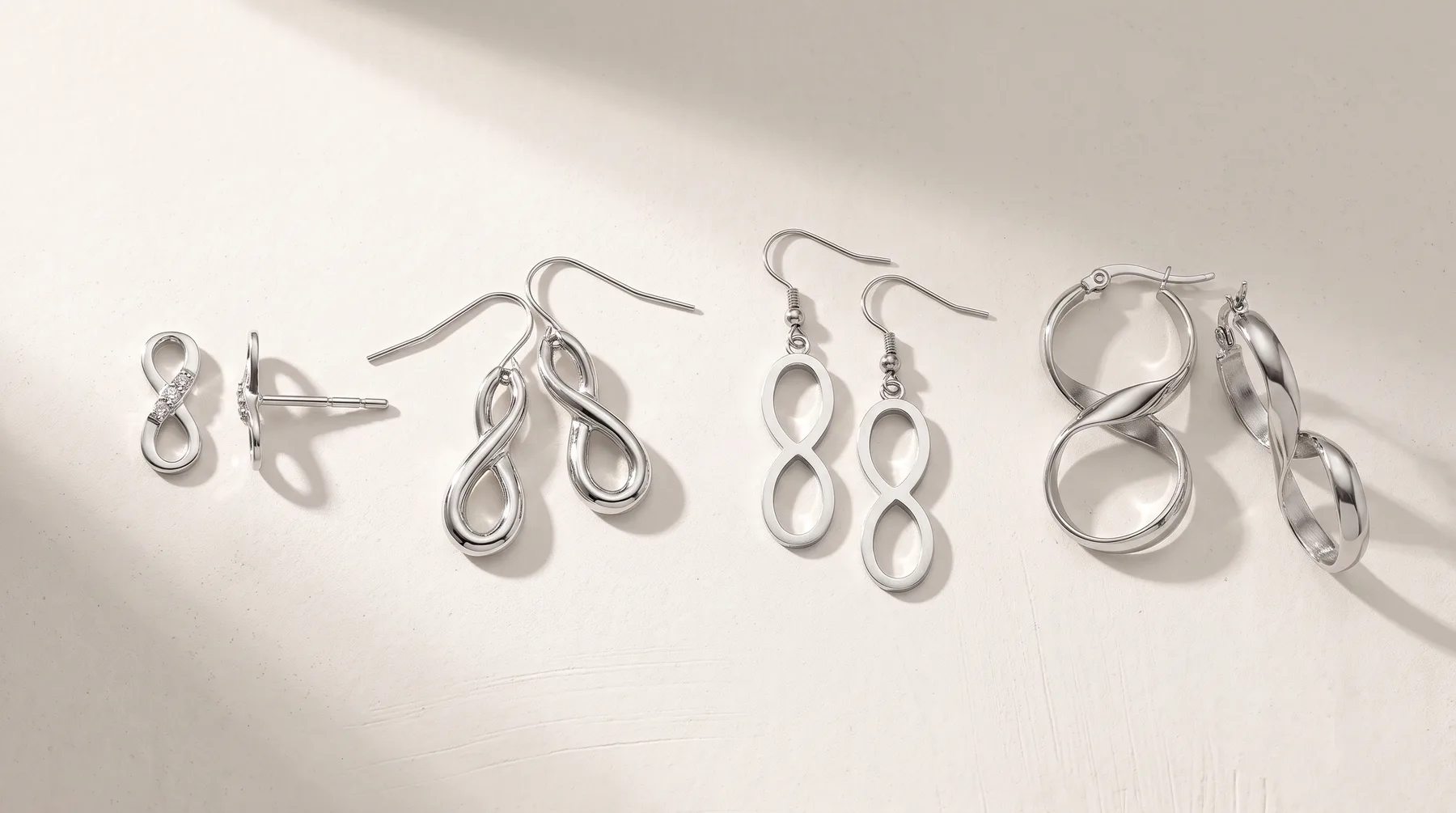Six pairs of silver earrings displayed on a white surface, including infinity symbol studs, twisted loop dangles, and polished hoop styles in various sizes.
