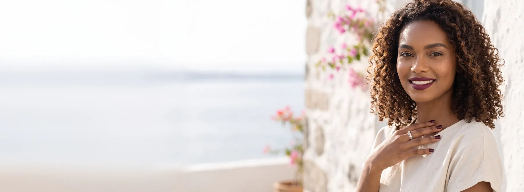 A smiling young woman with voluminous dark curls on a coastal balcony in soft morning light, her hand raised to her chest wearing a sterling silver intertwined hearts ring.