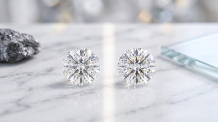 Two round brilliant-cut diamonds resting on a white marble surface, with a rough gray rock and glass panel blurred in the background.