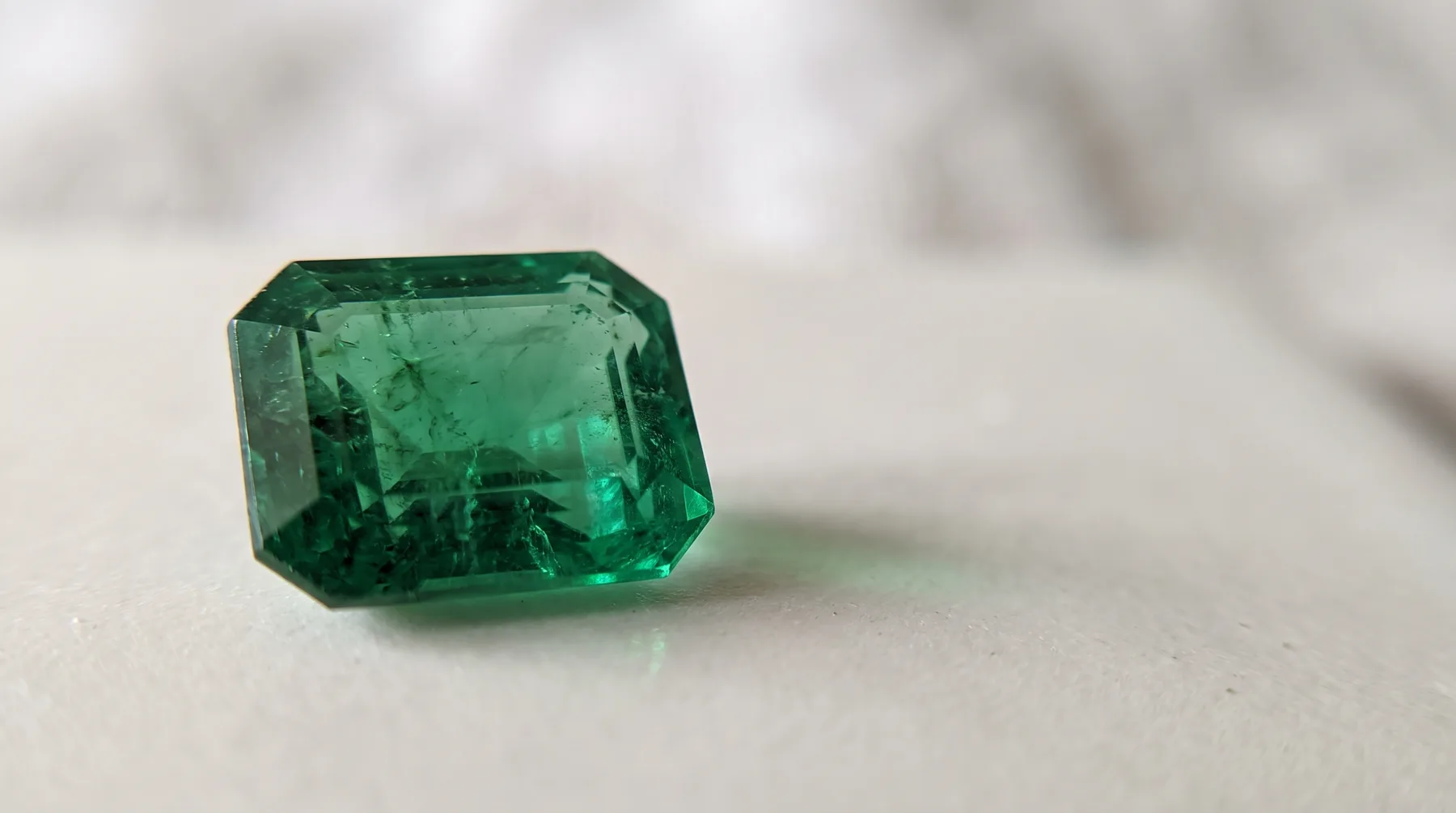 A rectangular step-cut emerald with cropped corners lying on a pale surface, its deep green interior showing the fine veiled inclusions jewelers call the jardin