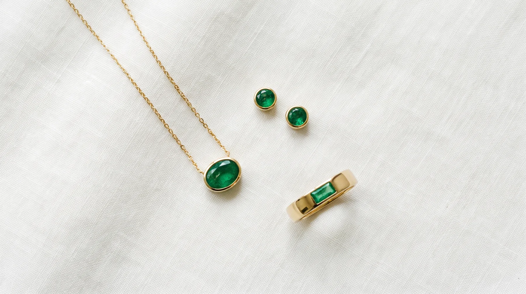 Three emerald pieces on ivory linen: a bezel-set oval emerald pendant on a fine gold chain, a pair of round emerald studs, and a gold band with a small rectangular emerald set flush into it