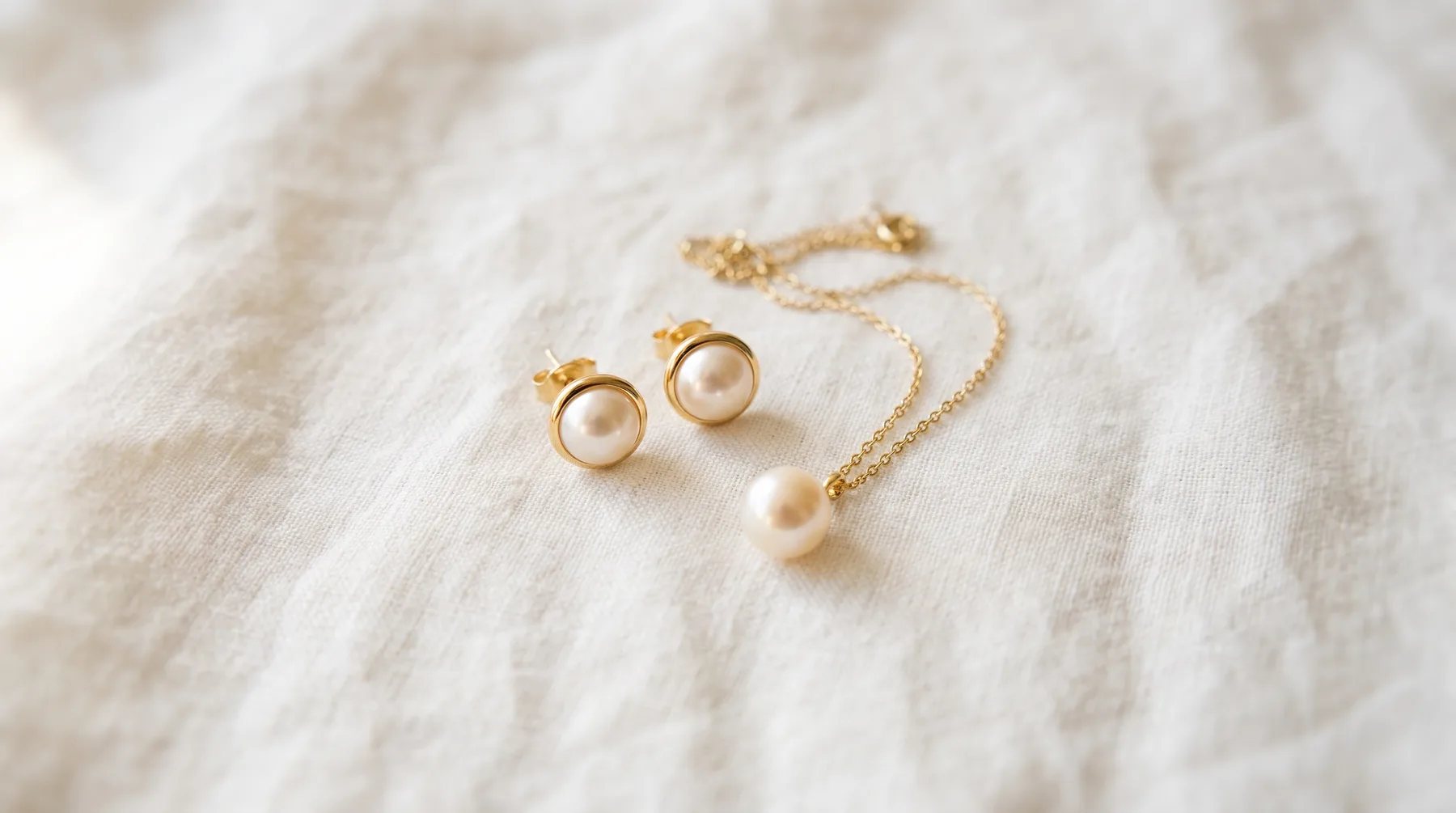 A pair of white pearl stud earrings in gold cups beside a single pearl hanging from a fine gold chain, laid on ivory linen