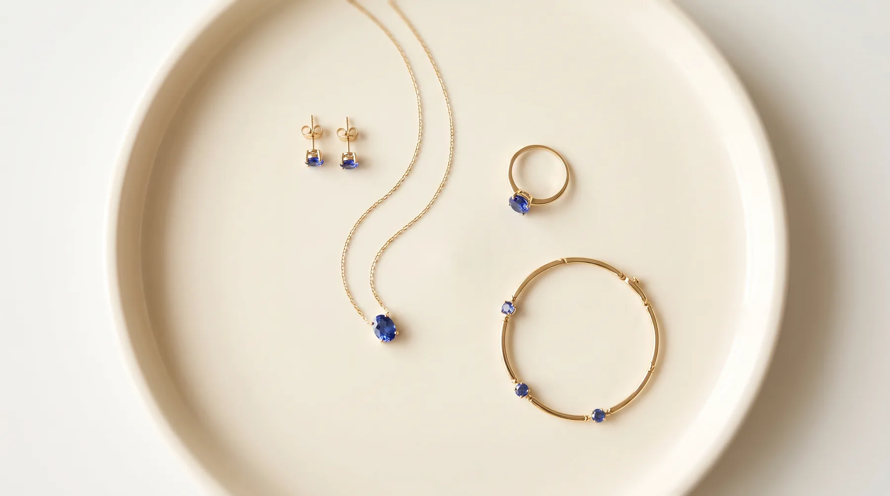 Four gold pieces set with blue sapphires arranged on a cream ceramic tray: a pair of studs, a fine chain with an oval pendant, a solitaire ring, and a slim bracelet with three round stones.