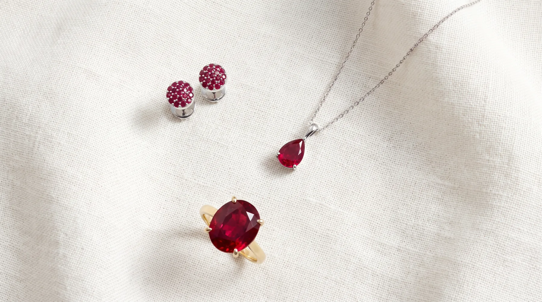 Three ruby pieces on ivory linen: white gold studs pave-set with small rubies, a pear-shaped ruby pendant on a fine white gold chain, and a yellow gold ring holding a large oval ruby in four prongs
