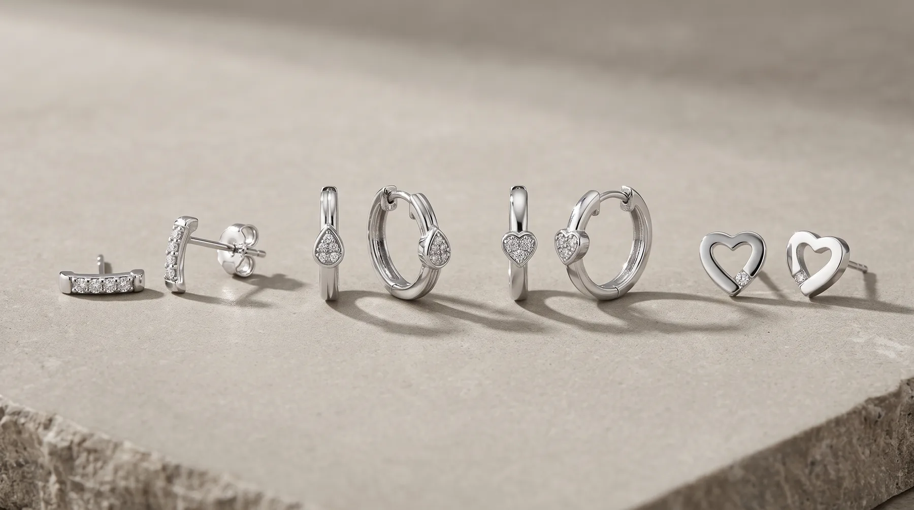 Seven silver jewelry pieces arranged in a line on a concrete surface, including earrings, hoops, and heart-shaped rings, all casting shadows.