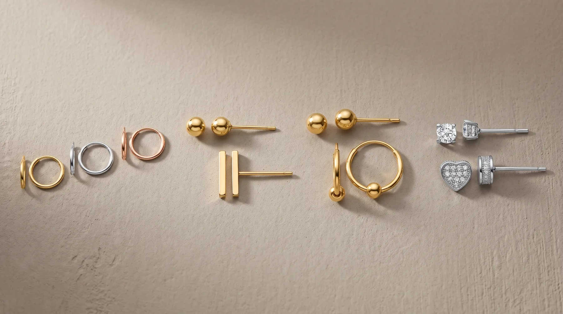 Six pairs of earrings arranged in a row on a beige surface, including gold and silver hoop earrings, ball studs, rectangular bar studs, and diamond stud earrings.