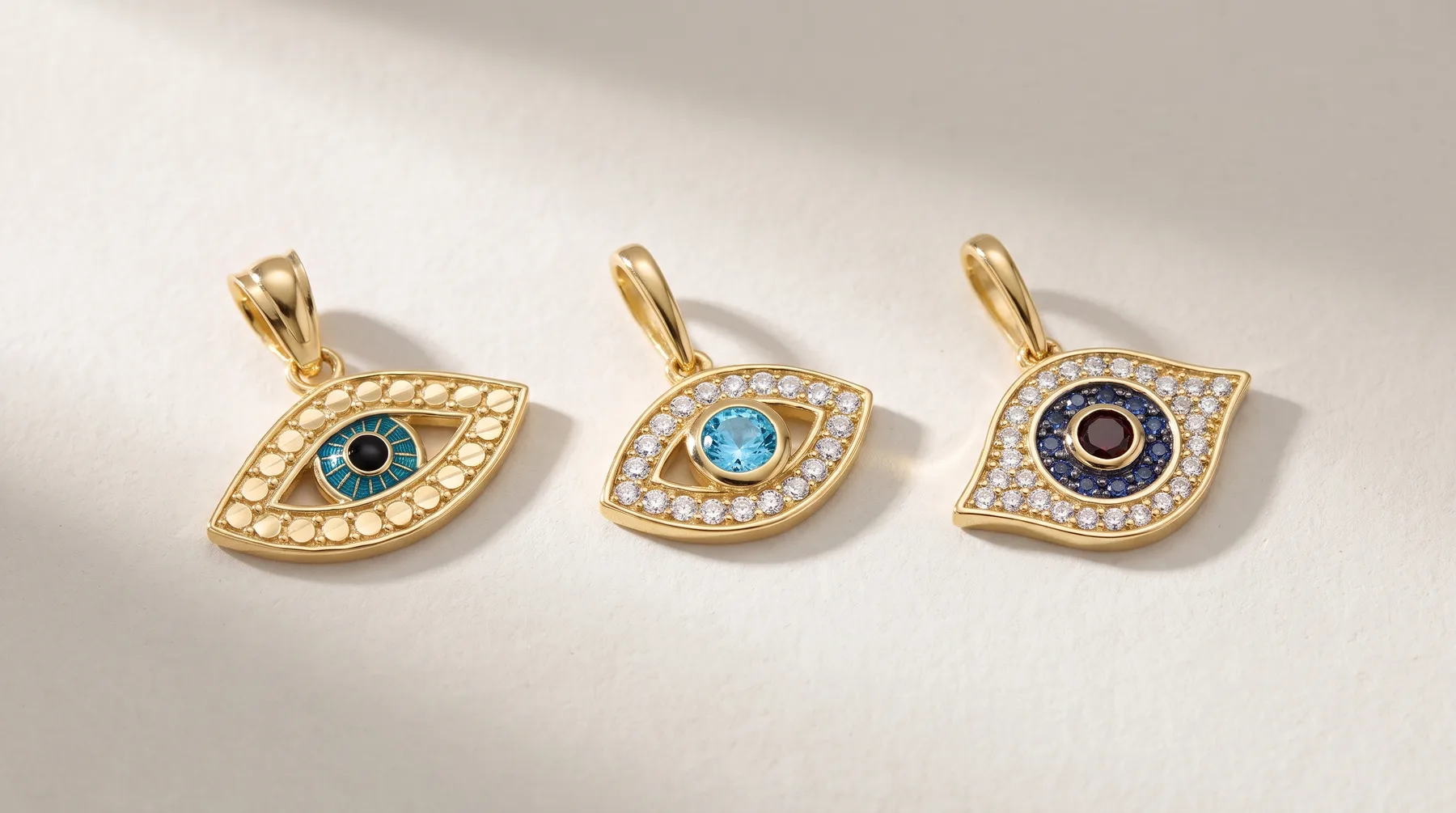 Three gold evil eye pendants with pearl and gemstone details, featuring turquoise, light blue, and dark blue center stones, arranged in a row on a white surface.