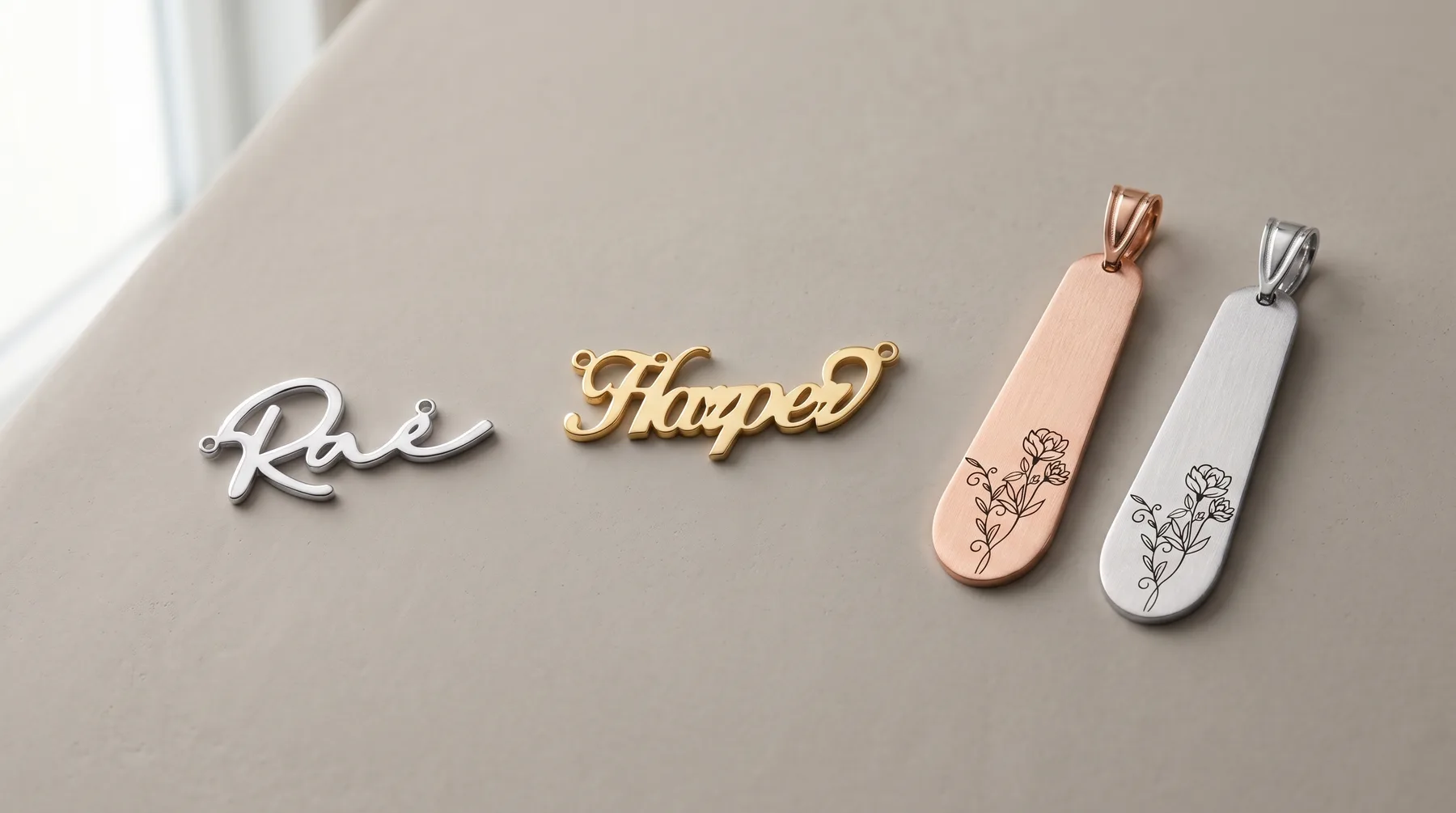 Four metal pendants on a beige surface: two script name charms in silver and gold, and two elongated oval pendants in rose gold and silver with delicate floral designs.