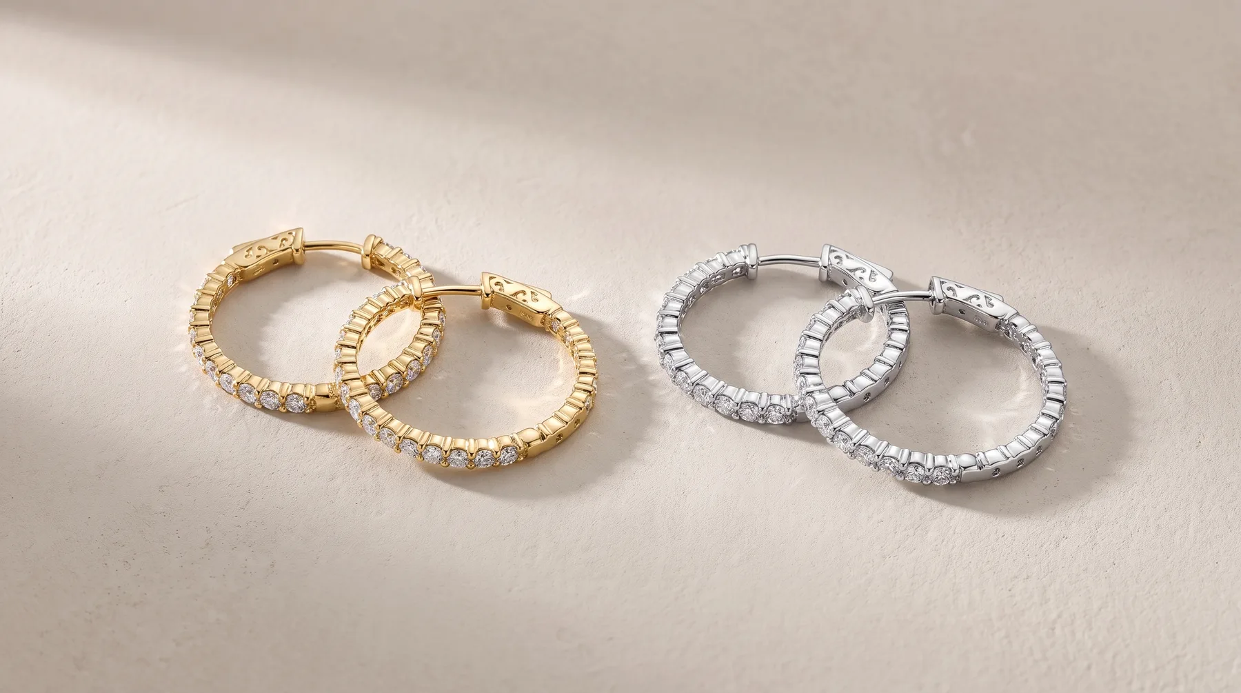 Two pairs of diamond hoop earrings displayed on a neutral surface: one pair in yellow gold and one pair in white gold, each with round diamonds set along the outer edge.