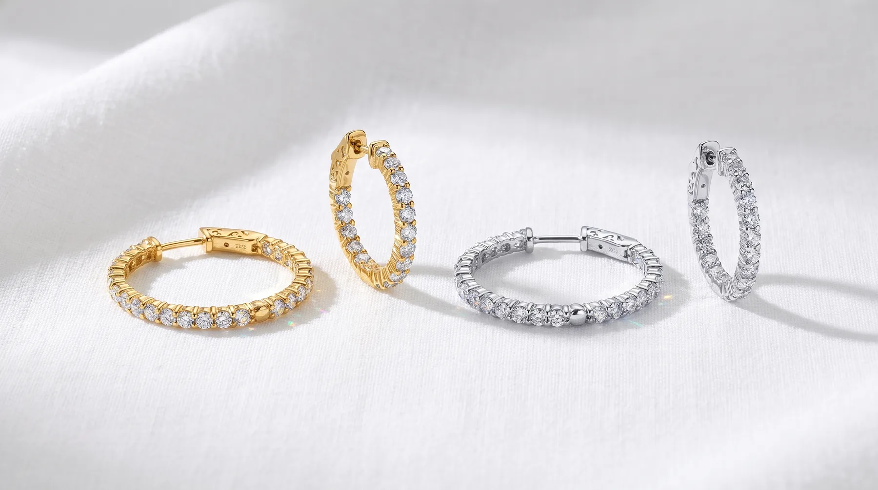 Two pairs of diamond hoop earrings displayed on a white surface, one pair in gold finish and one pair in silver finish, each with round brilliant-cut stones set along the curved band.
