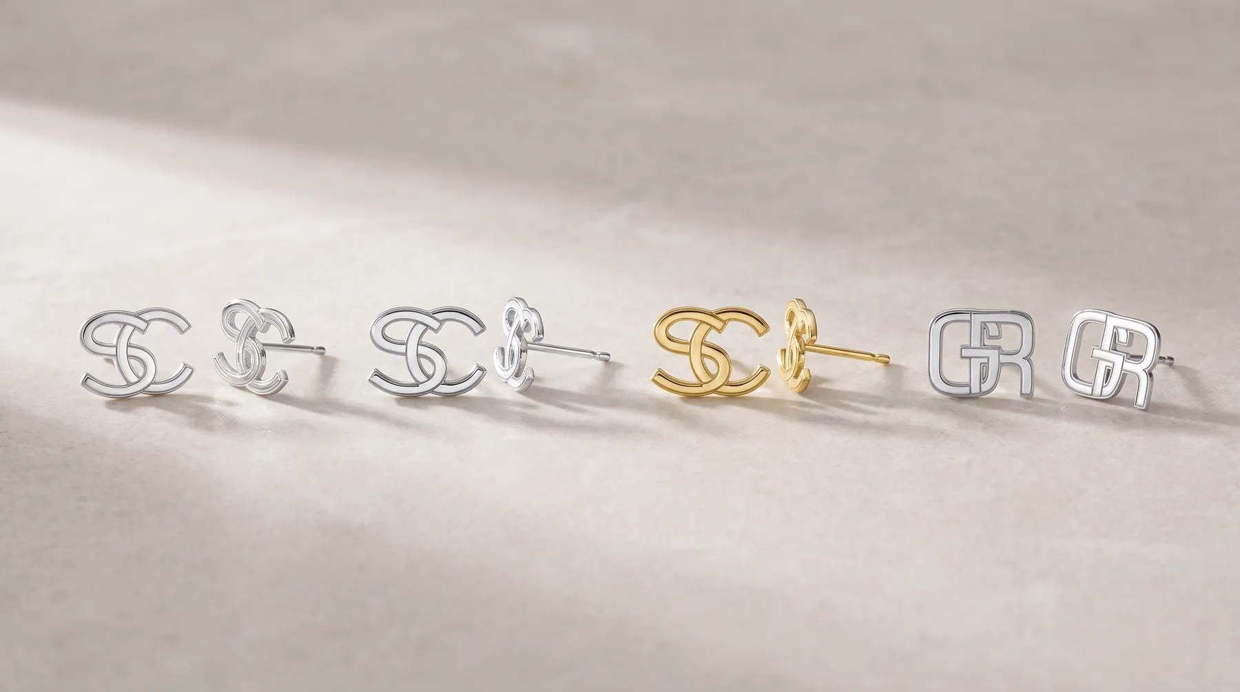 Six pairs of small stud earrings arranged in a row, including silver-toned and gold-toned interlocking letter designs and geometric shapes on a neutral textile surface.