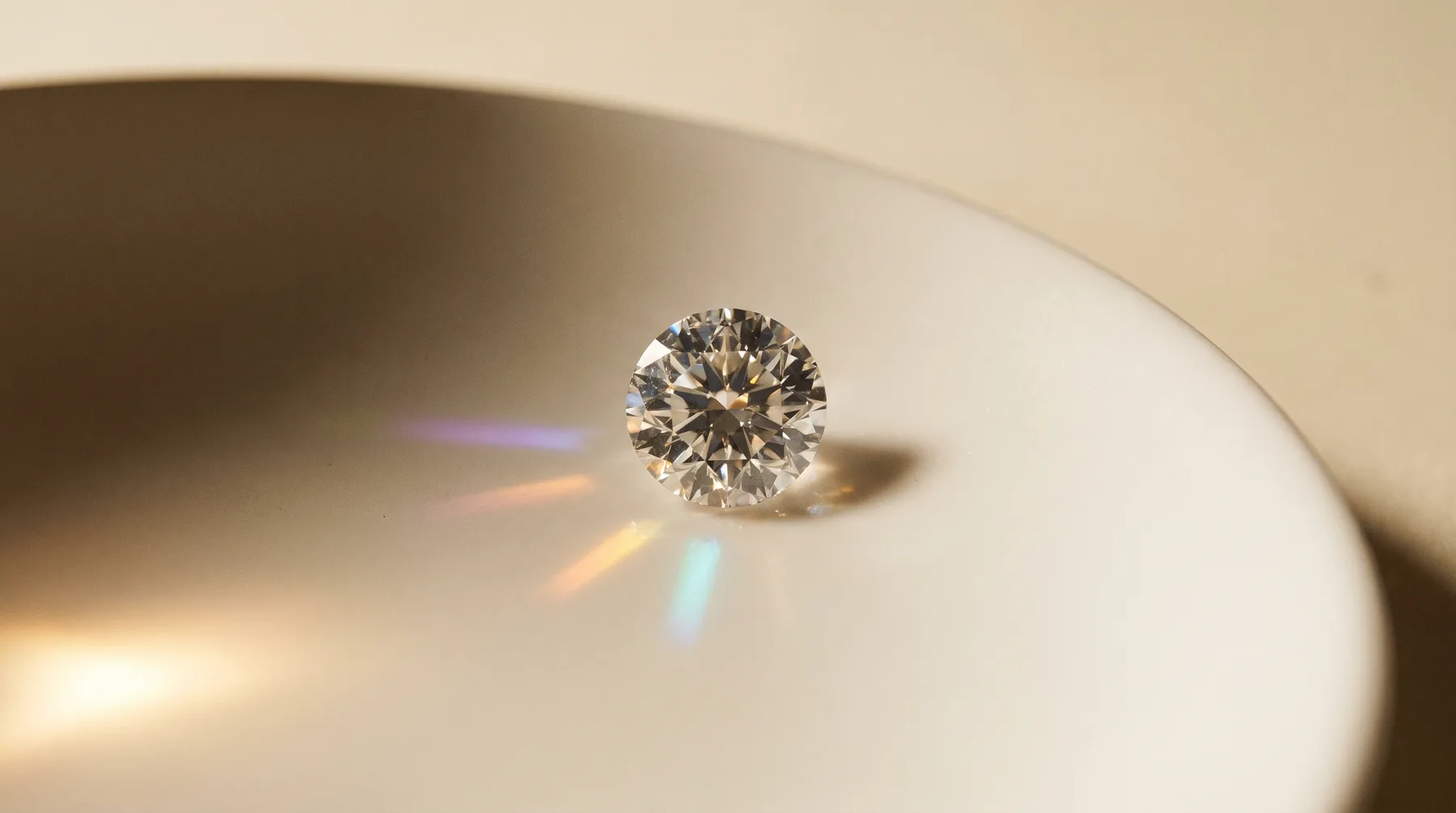 A single loose round brilliant diamond face-up on a pale porcelain dish, its facets showing a pattern of bright and dark areas while a side light throws small violet, orange and blue flashes across the glaze.