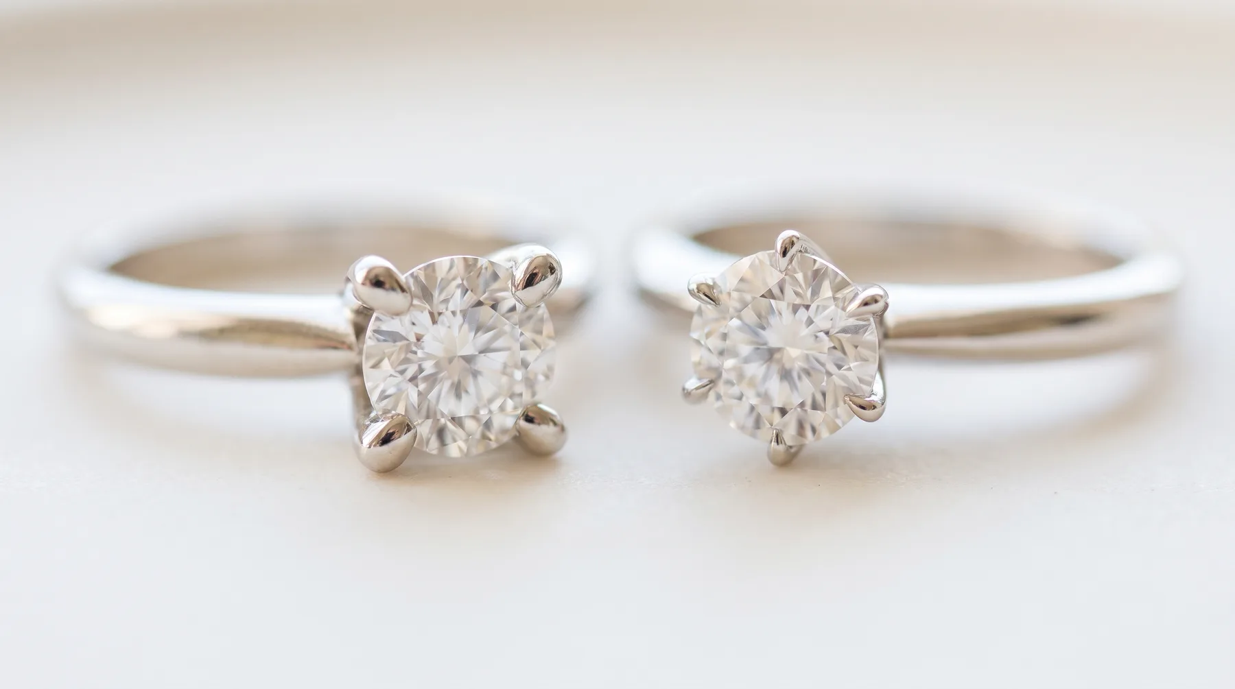 Two white metal solitaire rings side by side, the left holding a round diamond in four rounded prongs and the right in six slimmer claw prongs