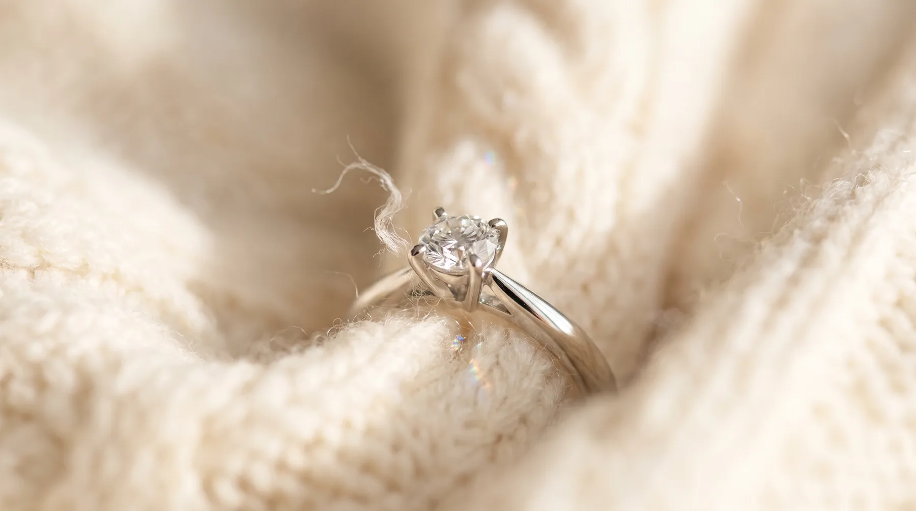 A white gold four-prong diamond solitaire nestled in a cream cable-knit sweater, a loose wisp of yarn caught near one prong